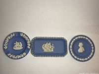 Three Wedgwood jasperware plates: two round with white figure reliefs and wreath borders, one rectangular with classical white relief scene and border.
