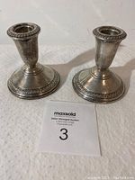 Front view of two silver candlesticks showing decorative rim and base detail on a white surface with auction number card 3 in front