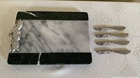 Photo showing the rectangular marble cheeseboard with white and dark green marble sections, and the set of four pâté spreaders with bunny designs on handles arranged beside it.