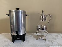 Photo showing a large cylindrical stainless steel coffee urn with black plastic side handles and a spout, standing next to a decorative silver-tone metal and glass coffee warmer with a lid and handle on a tripod stand.