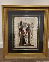 Framed Ancient Egyptian papyrus art with hieroglyphics and two standing female figures, gold frame with black mat.