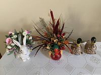 Photo showing the three centrepieces on a table: glass rabbit vase with flowers, papier-mache duck with flowers, and metal duck pair.