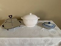White ceramic soup tureen, mid-century 2-tiered serving plate, and blue-white enamelware casserole dish all displayed on a patterned white tablecloth.