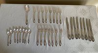 Full set of flatware items including 8 knives, 7 dinner forks, 8 salad forks, 8 spoons, and 1 large serving spoon arranged on tablecloth.