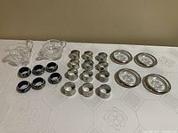 Full lot overview showing cream and sugar set, pewter napkin rings, painted wooden napkin rings, and crystal silver coasters arranged on table.