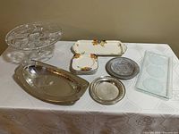 Wide shot of all items on tablecloth: pedestal crystal cake plate, floral serving sandwich plate, six small square plates, two round wine coasters, silver plated bread basket.