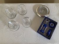 Four clear stemless wine glasses, metal wine coaster, and boxed blue corkscrew set placed on tablecloth.