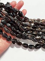 Close-up of multiple strands of tumbled smoky quartz beads held in hand, showing shape, color, and natural blemishes