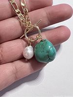 Close-up of the chunky turquoise and pearl pendant hanging from a circular ring on a gold plated chain held in hand