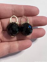 Pair of earrings held in hand showing large faceted black onyx stones on gold plated hoops.