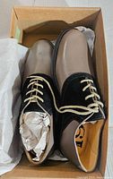 Pair of men's leather oxford shoes in box with paper stuffing inside, highlighting tan pebble leather body, black suede accents, cream laces, and brand insoles
