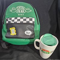 Green Central Perk backpack with checkerboard front pocket and enamel pins next to white Central Perk ceramic travel mug with green lid