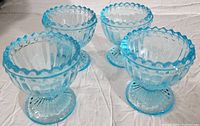 Four blue glass egg holders with scalloped rims and pedestal bases placed together on white fabric.