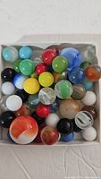 Top view of a box containing about 37 colorful vintage glass marbles with different patterns and sizes including one larger shooter marble.