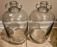 Pair of large, heavy, clear glass jugs standing side by side on tile floor against wall, showing overall shape and size.