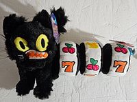 Two Bark dog toys on a white surface, a black cat plush toy and a slot machine roller toy with colorful embroidered symbols. Both with tags.