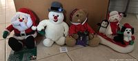 Four plush animated Christmas characters: Santa, Frosty the Snowman, a teddy bear with a book, and a trio on a sled featuring a penguin, polar bear, and dog.