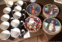 Photo showing ten tall Christmas-themed ceramic mugs and 28 plates with festive illustrations, arranged on a kitchen counter.