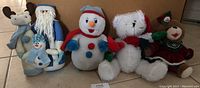 Front view of four Christmas stuffed decorations arranged in a row on floor: moose with snowman, blue Santa, snowman, white teddy bear, and dressed reindeer.