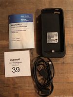 Photo showing the black Bauhn charging case for iPhone 5/5S alongside a printed user manual and a USB charging cord on a wood surface.