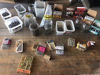 Wide view of table with multiple containers of assorted nails, screws in jars, plastic tubs, cardboard boxes, and bags.