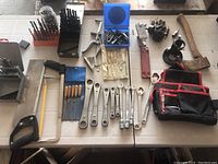 Top-down photo of assorted hand tools including drill bits, wrenches, saw frame, files, sockets, and a hatchet axe.