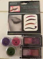 Photo including the adhesive eyeliner pack along with three shimmer powders in purple, pink, and green, and two dual-color eye shadow compacts with shades of pink and purple.