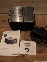 Philips small bedside alarm clock with power adapter and user manual laid out on wooden surface. Clock is black with digital display and Philips branding on top.