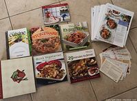 Photo of five Pampered Chef cookbooks, a recipe collection binder, and a large stack of recipe pages and cards spread out on the floor.