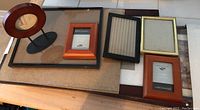 Photo showing a group of 9 assorted photo frames laid flat on a surface with various sizes and styles including round wooden frame, small reddish-brown frames, and medium to large plain wood or black bordered frames.
