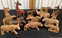 Overview of fourteen carved wood African animal figures and napkin rings including giraffes, elephants, leopards, zebra, and other animals arranged on black surface.