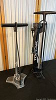 Two manual tire pumps side by side on black surface against wood and metal background.