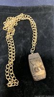 Full view of the gold-tone chain and attached Aztec style rectangular pendant on black background.