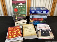 Photo shows a stack and scattered arrangement of various books including 'Atlas Shrugged' by Ayn Rand, 'Countdown to Socialism' by Devin Nunes, and others, placed on a wooden table against a curtain backdrop.