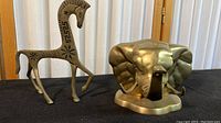 Photo showing brass stylized horse figurine with decorative engravings and elephant head brass figurine side by side on black surface.