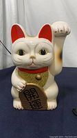 Front view of white ceramic Lucky Cat statuette with raised right paw, red ears and collar, gold glitter bib, and kanji plaque