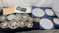 Overview of all vintage tablecloths and doilies on dark fabric background, showing cloth tablecloths, linked crocheted doilies, and packaged paper doilies