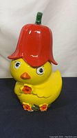 Front view of the yellow ceramic duck cookie jar with red bell pepper lid showing details of the face, flowers, and overall condition.