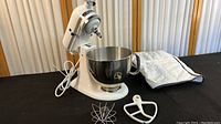 White KitchenAid stand mixer with attached stainless steel mixing bowl, two mixer attachments (a wire whisk and a flat beater), and a protective fabric cover beside it.