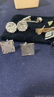 Photo showing round rhinestone cufflinks, square star-engraved cufflinks, gold-tone tie clip, triangular pin, and other pins on a black cloth background.