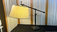 Metal adjustable table lamp with black base, long adjustable arm, and beige fabric shade on black cloth background in front of a light curtain