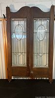 Front view of both wooden swinging doors with ornate glass panels clearly showing oval and geometric leaded glass design and wood finish.