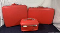 Three red vintage American Tourist suitcases arranged with the small suitcase in front and two larger ones behind, shown on a black surface with white fabric background.