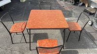 Top view of the square vintage Cosco folding table with four matching metal folding chairs with orange patterned cushioned seats arranged around it on an asphalt surface.