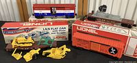 Four Lionel model train cars shown with respective original boxes: Illinois Central boxcar, L&N flat car with bulldozer and scraper models, US Mail car, and an operating searchlight car. All cars appear well preserved and boxed.