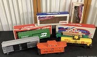 Five vintage Lionel train cars laid out in front of their boxes including Shell Oil tanker, log dump car, Illinois Central caboose, Burlington Northern boxcar, and Baltimore & Ohio boxcar.