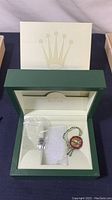 Green Rolex watch box open to show cream interior lining, plastic bag with extra links and Rolex tag hanging inside.