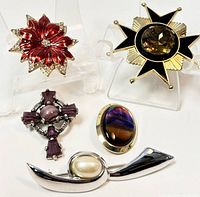 Five vintage brooches displayed on clear holders including red enamel floral, black star, purple geometric, oval cabochon, and silver faux pearl designs.