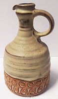 Front view of the pottery jug showing the round handle, beige glaze, and carved terracotta base with decorative symbols.