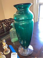 Full view of the taller green Blenko vase with a wide base, sitting on a wooden surface next to a greenish glass bottle and small figurines.